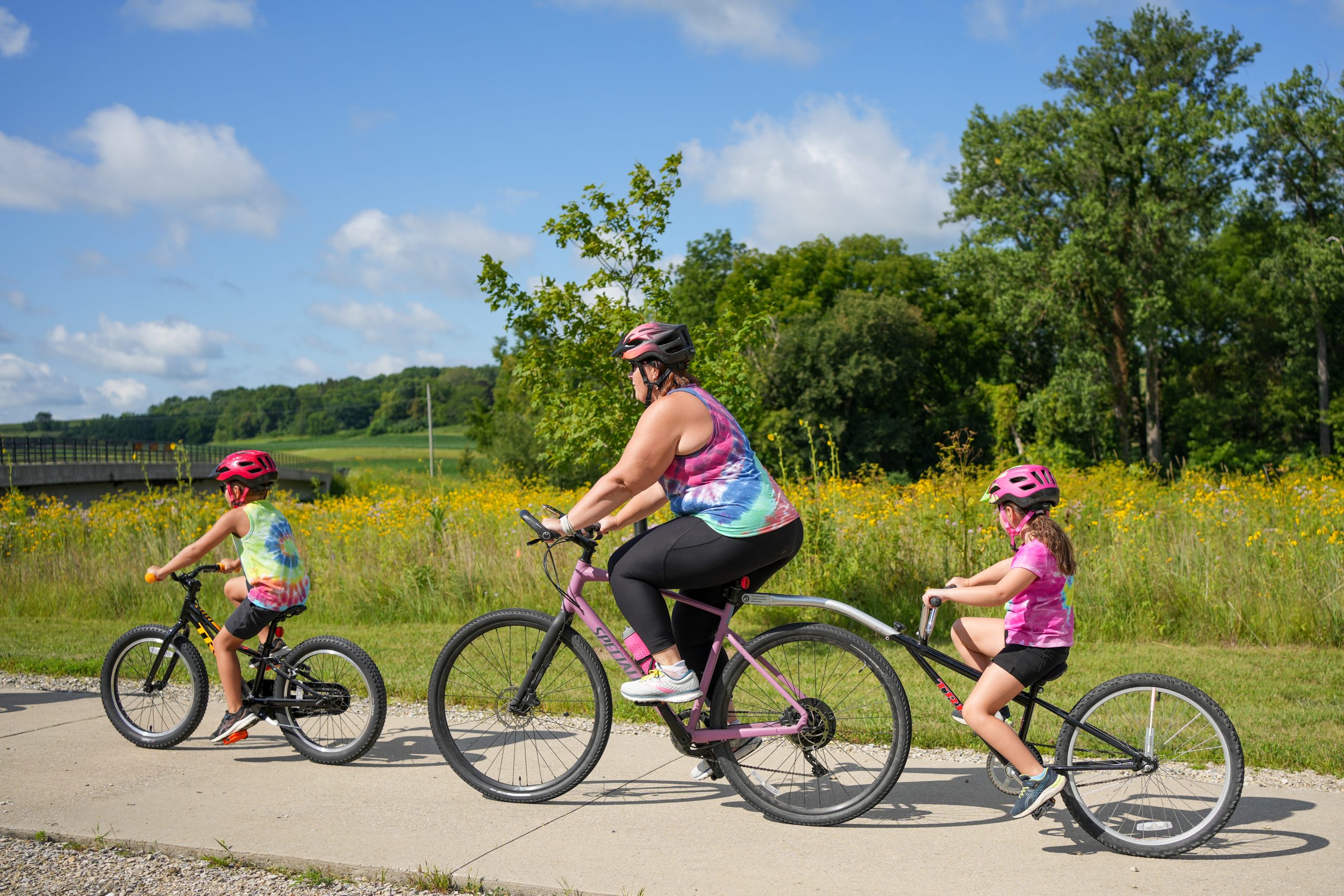 Two kids and an adult riding bikes on a concrete trail against a blue sky background.