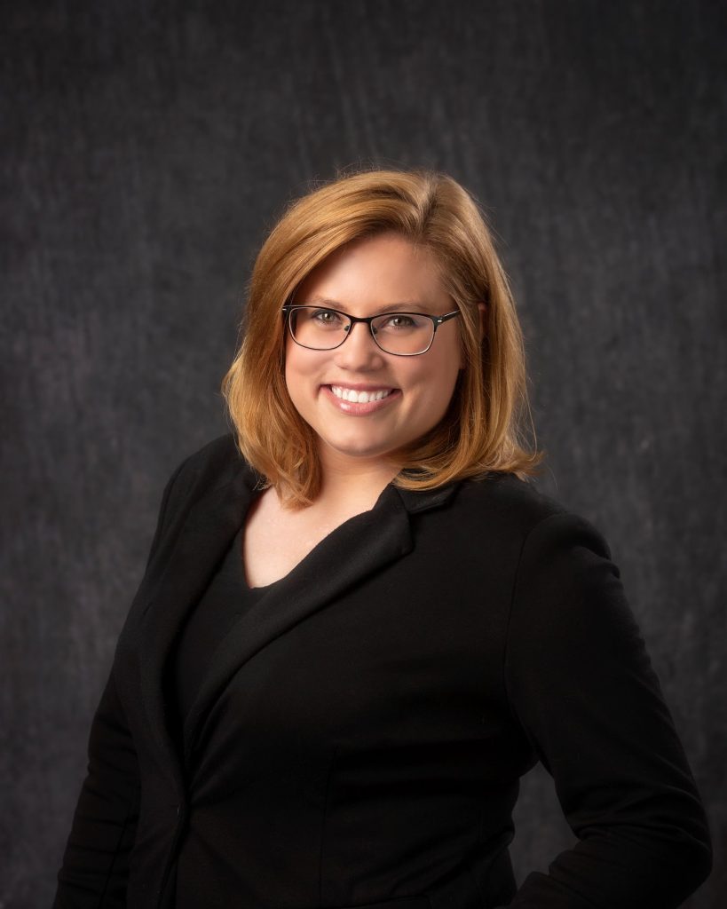 Woman with strawberry blonde hair smiling and wearing glasses and a black blazer.